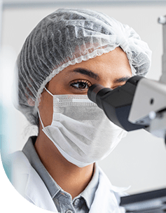 Nahaufnahme einer Ärztin mit Schutzhaube und Gesichtsmaske, die ein Mikroskop in einem Labor benutzt.