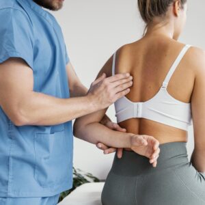 Ein männlicher Osteopath in blauer Berufskleidung überprüft den Schulterknochen einer weiblichen Patientin. Die Patientin sitzt und trägt ein weißes Sport-BH und graue Leggings. Der Osteopath tastet die Schulter der Patientin mit beiden Händen ab.
