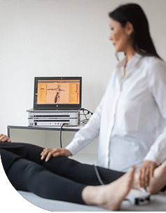 Eine Ärztin führt eine Vitalfeldmessung an den Beinen einer liegenden Patientin durch. Sie benutzt ein modernes Gerät mit einem Bildschirm, auf dem ein menschlicher Körper dargestellt ist.