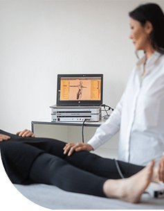 Therapeutin führt eine Vitalfelddiagnostik an einer Patientin durch, deren Beine auf einer Behandlungsliege ausgestreckt sind, mit einem Bildschirm im Hintergrund, der einen schematischen Körper zeigt.