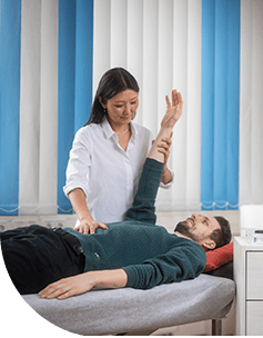 Therapeutin korrigiert die Armhaltung eines auf dem Rücken liegenden Patienten in einer Praxis mit hellblauen Vorhängen im Hintergrund