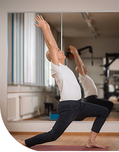 Ein Mann in Weiß führt eine anspruchsvolle Yoga-Pose aus, bei der er sich rückwärts beugt, während sein Bein gestreckt ist, in einem hellen Raum mit Spiegelwand