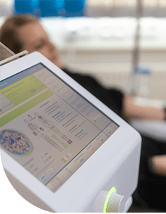 Nahaufnahme eines modernen medizinischen Geräts mit einem Bildschirm, der verschiedene gesundheitsbezogene Statistiken anzeigt, im Hintergrund eine unscharf sichtbare, entspannte Person.