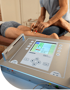 Ein modernes Mikrostromtherapiegerät mit einem Touchscreen, der verschiedene Einstellungen anzeigt, während ein Therapeut die Behandlung an einem Patienten, der auf einer Liege sitzt, durchführt.